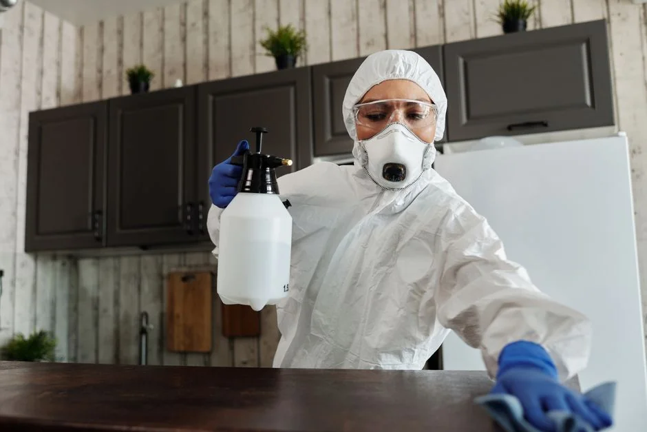 Person in protective suit disinfecting a white table indoors with a sprayer.